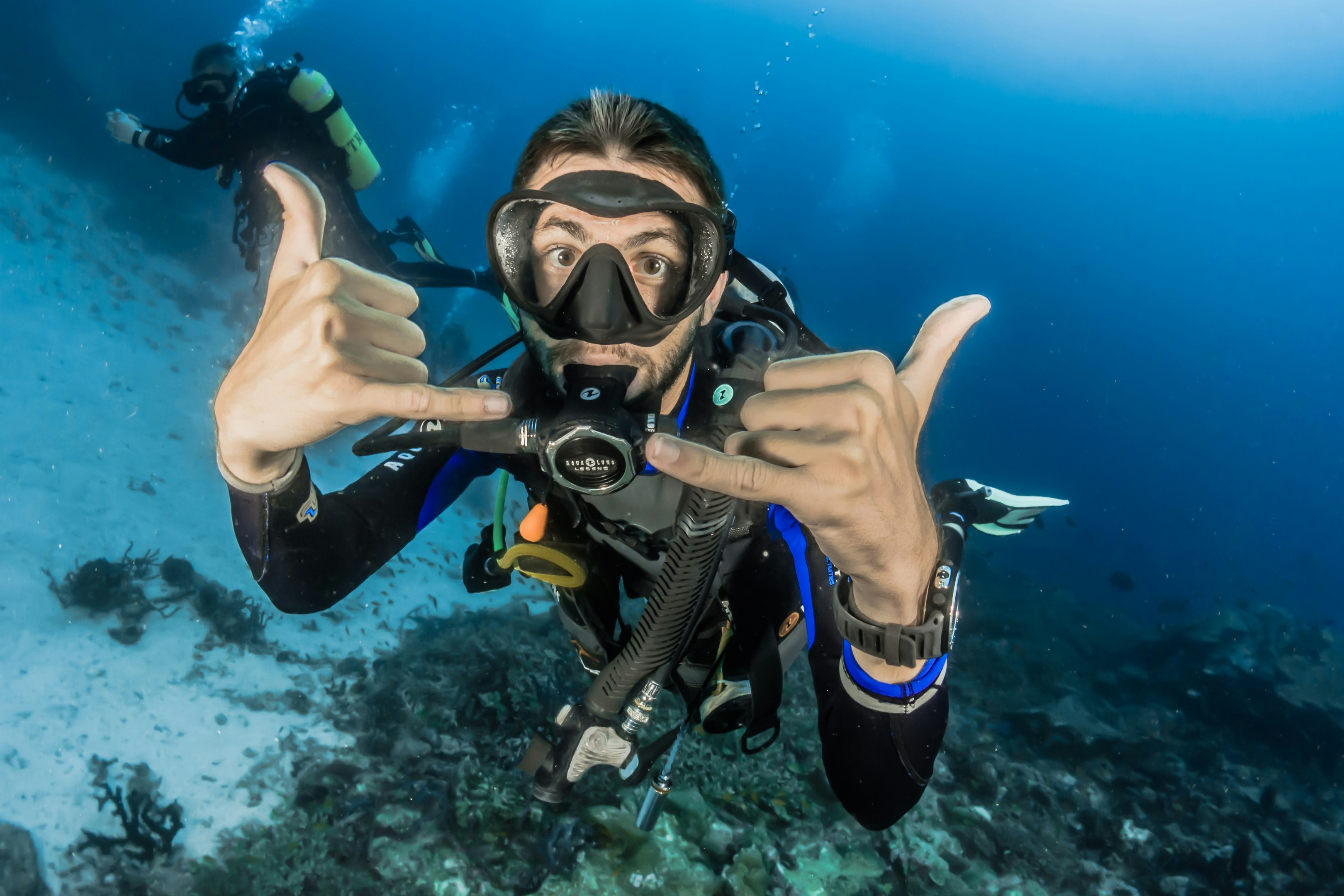 Ein technischer Sidemount-Taucher steigt durch klares blaues Meerwasser zur Bootshülle auf. Sonnenlicht durchdringt die Oberfläche und erzeugt helle Muster über dem Taucher. Der Taucher trägt komplette Ausrüstung.